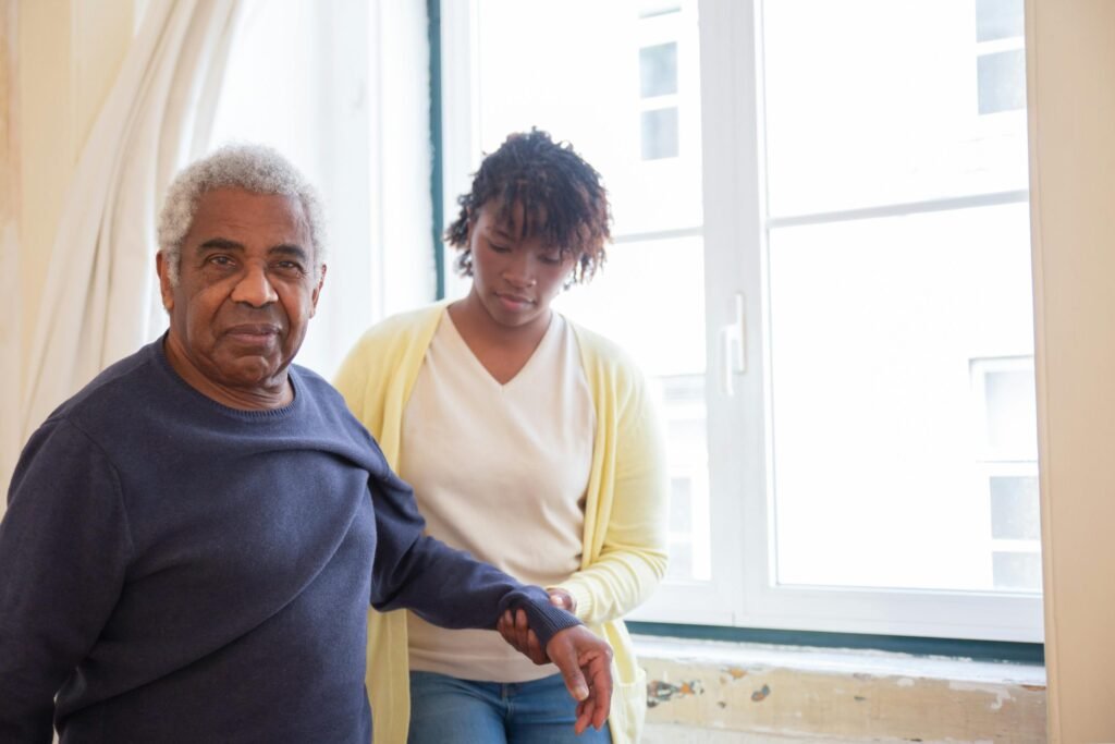 An old man with his caregiver by the window giving him support